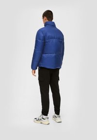 Blauwe gewatteerde jas met een hoge kraag, gedragen over zwarte cargobroeken en sneakers, bekeken vanaf de achterkant. Het materiaal lijkt glanzend en geïsoleerd.