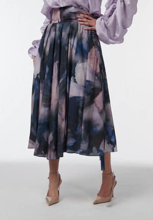 Personne portant une jupe midi plissée à motifs aquarelle, un chemisier lavande à manches bouffantes et des escarpins nude, debout une main sur la hanche.