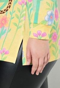 Żółta kwiatowa bluzka z różowo-niebieskim wzorem kwiatowym oraz bocznym rozcięciem. Połączona z błyszczącymi czarnymi legginsami.