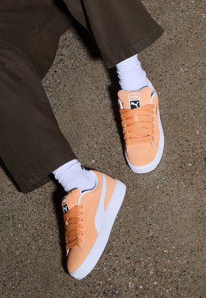 Personne portant des baskets Puma couleur pêche avec des semelles blanches, des chaussettes blanches et un pantalon marron foncé, assise sur un sol en béton tacheté.