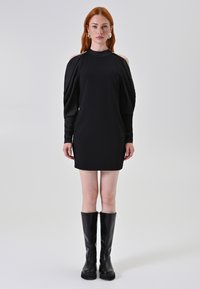 Schwarzes kurzes Kleid mit langen, lockeren Ärmeln und Ausschnitten an den Schultern. Getragen mit schwarzen kniehohen Stiefeln, die eine schlanke, moderne Silhouette kreieren.