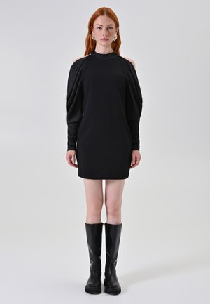 Schwarzes kurzes Kleid mit langen, lockeren Ärmeln und Ausschnitten an den Schultern. Getragen mit schwarzen kniehohen Stiefeln, die eine schlanke, moderne Silhouette kreieren.