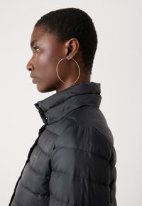 Black puffer jacket with a high collar and a quilted design. Features a smooth texture and a close-fitting silhouette. Silver hoop earrings are visible.