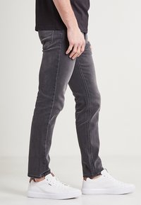 Jeans de corte slim para hombre en denim gris oscuro, con un clásico diseño de cinco bolsillos y una sutil textura desvanecida. Combinados con zapatillas blancas.