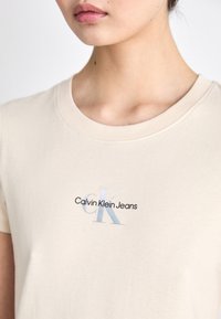 Béžové bavlnené tričko s okrúhlym výstrihom, na ktorom je potlačené logo v modrom a čiernom texte s nápisom "Calvin Klein Jeans."