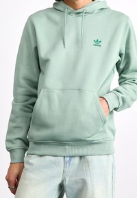 Mintgrön luvtröja i mjukt material, med en ficka framtill och broderad logotyp på bröstet, samt ribbade muddar och fåll.