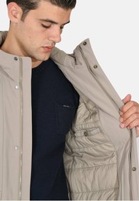 Giacca esterna beige con chiusura a zip e bottoni automatici, che mostra un maglione blu navy texturizzato sotto; fodera imbottita interna.