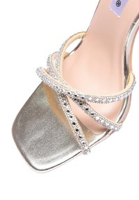 Sandalo inargento metallico con cinturini, impreziosito da decorazioni simili a diamanti, punta quadrata e una morbida soletta color nude. I cinturini si intrecciano in modo elegante.