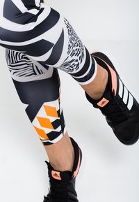 Legginsy w czarno-białe wzory mają geometryczne, zwierzęce i abstrakcyjne motywy z pomarańczowymi akcentami, w połączeniu z czarnymi butami sportowymi.