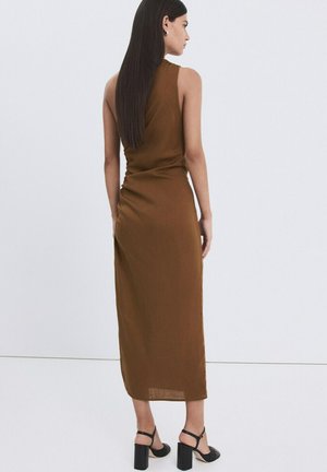 Mujer de cabello largo y oscuro, vestida con un vestido marrón sin mangas que llega hasta los tobillos y sandalias negras de tacón alto, de pie contra un fondo sencillo.