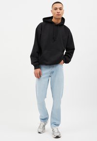 Sudadera de algodón negra con un bolsillo frontal, combinada con jeans azul claro y zapatillas plateadas. La sudadera tiene un cordón y un ajuste oversize.