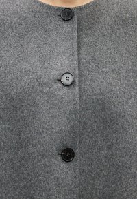 Hall hallist fliiskangast mantel, millel on ümmargune kaelus, kolm musta nööd, ning sile eesmine osa ilma nähtavate kinnituste või mustriteta.