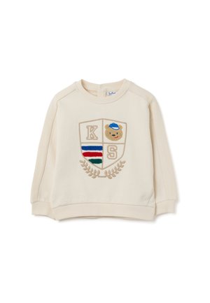 Sweatshirt crème avec des poignets et un ourlet côtelés, comportant un patch brodé en forme de bouclier avec les lettres "K", "S" et un design de teddy bear.