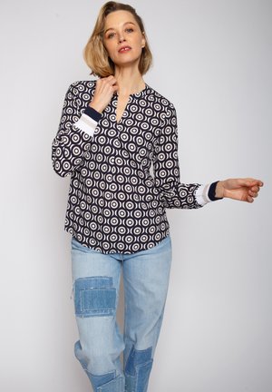 Marineblaue Bluse mit kreisförmigen weißen Mustern, ausgestattet mit einem V-Ausschnitt und langen Ärmeln mit gestreiften marineblauen und weißen Bündchen. Kombiniert mit hellblauen, stark abgenutzten Jeans.