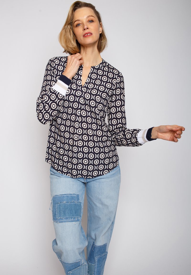 Marineblaue Bluse mit kreisförmigen weißen Mustern, ausgestattet mit einem V-Ausschnitt und langen Ärmeln mit gestreiften marineblauen und weißen Bündchen. Kombiniert mit hellblauen, stark abgenutzten Jeans.