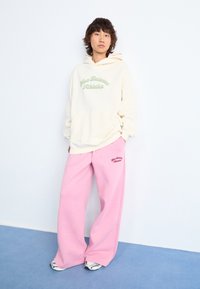 Personne se tenant contre un mur blanc portant un sweat à capuche crème et un pantalon de jogging rose à jambes larges avec des baskets sur un sol bleu.