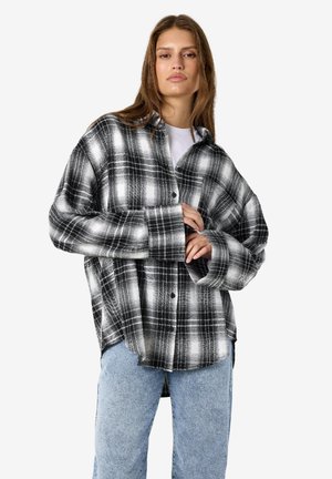 Ung kvinde iført en oversized sort-hvid ternet skjorte med lyseblå jeans mod en ensfarvet baggrund.