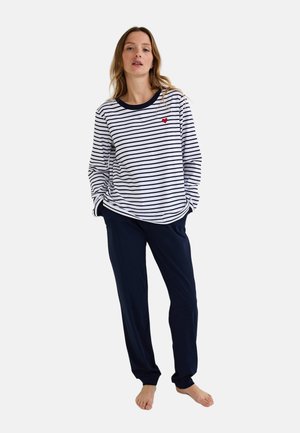 Femme debout pieds nus portant un haut à manches longues blanc et bleu marine rayé avec un petit cœur rouge et un pantalon bleu marine.