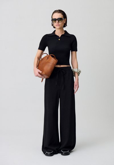 Polo shirt nero a righe con maniche corte e un bottone dorato, abbinato a pantaloni neri a gamba larga. Tiene una borsa a secchiello in pelle marrone.