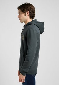 Hoodie verde scuro con cappuccio regolabile, tasca frontale e polsini a coste. Presenta un logo dorato sul petto. Texture liscia e design casual.