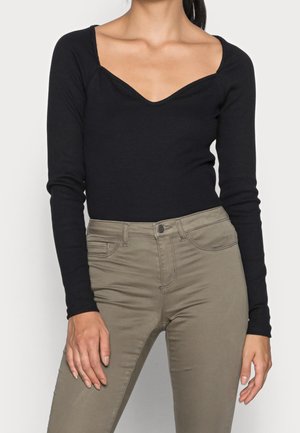 Femme portant un haut noir à manches longues avec un décolleté en cœur et un pantalon beige taille haute, les mains reposant sur les hanches.