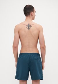 Tengerés kék úszóshort könnyű anyagból, rugalmas derékpánttal és hátsó zsebbel, egy mezítelen háttal rendelkező modellen bemutatva.