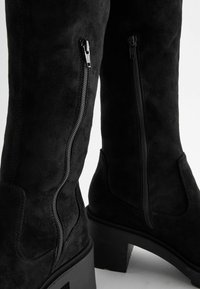 Gros plan sur des bottes montantes en daim noir, avec deux fermetures éclair verticales sur les côtés et des talons carrés épais.