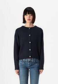 Navy blauwe gebreide cardigan met een ronde halslijn, voorzien van zilveren hartvormige knopen en lange mouwen, gecombineerd met lichtblauwe jeans.