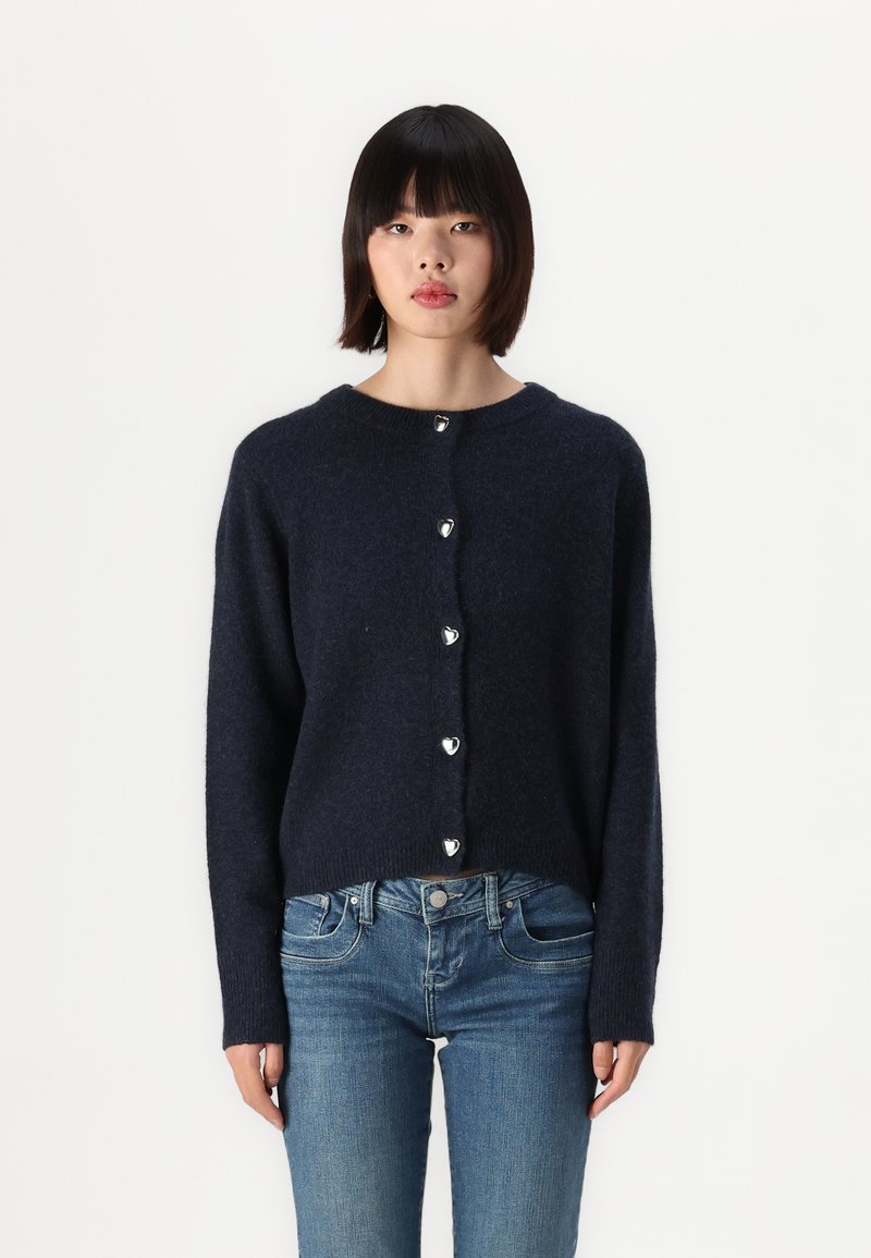 Navy blauwe gebreide cardigan met een ronde halslijn, voorzien van zilveren hartvormige knopen en lange mouwen, gecombineerd met lichtblauwe jeans.