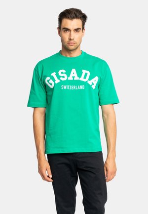 Mann trägt ein hellgrünes kurzärmliges Hemd mit dem Text "Gisada Switzerland", kombiniert mit schwarzer Hose, steht vor einem schlichten Hintergrund.