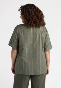 Camisa verde oliva de manga corta, con rayas verticales y patrones texturizados. Corte holgado con cuello redondo y tejido suave.