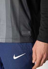 Atletické tričko s čiernymi a sivými pruhmi, s etiketou označenou ako "Navrhnuté." V kombinácii s tmavými modrými textúrovanými nohavicami a bielym logom Nike.