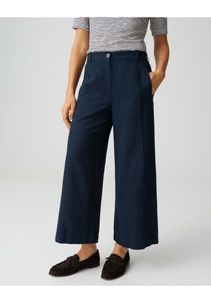 Personne portant un pantalon large cropped bleu marine, des mocassins noirs avec des nœuds, et un haut à manches courtes rayé, debout avec une main dans la poche.