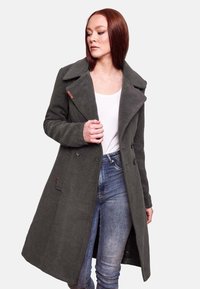 Manteau en laine vert foncé avec un large col, des boutons double boutonnage et des poches latérales, porté sur un haut blanc et un jean bleu.