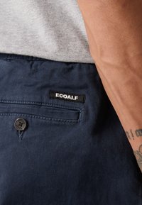 Nahaufnahme von einer Tasche in marineblau mit ECOALF-Label und Knopf, getragen von einer Person in hellgrauem Shirt, die einen tätowierten Arm zeigt.