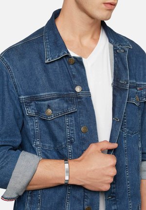 Mand iført en blå denimjakke over en hvid V-hals t-shirt, holder jakken ved kraven med den ene hånd og bærer et sølv- og sort armbånd.