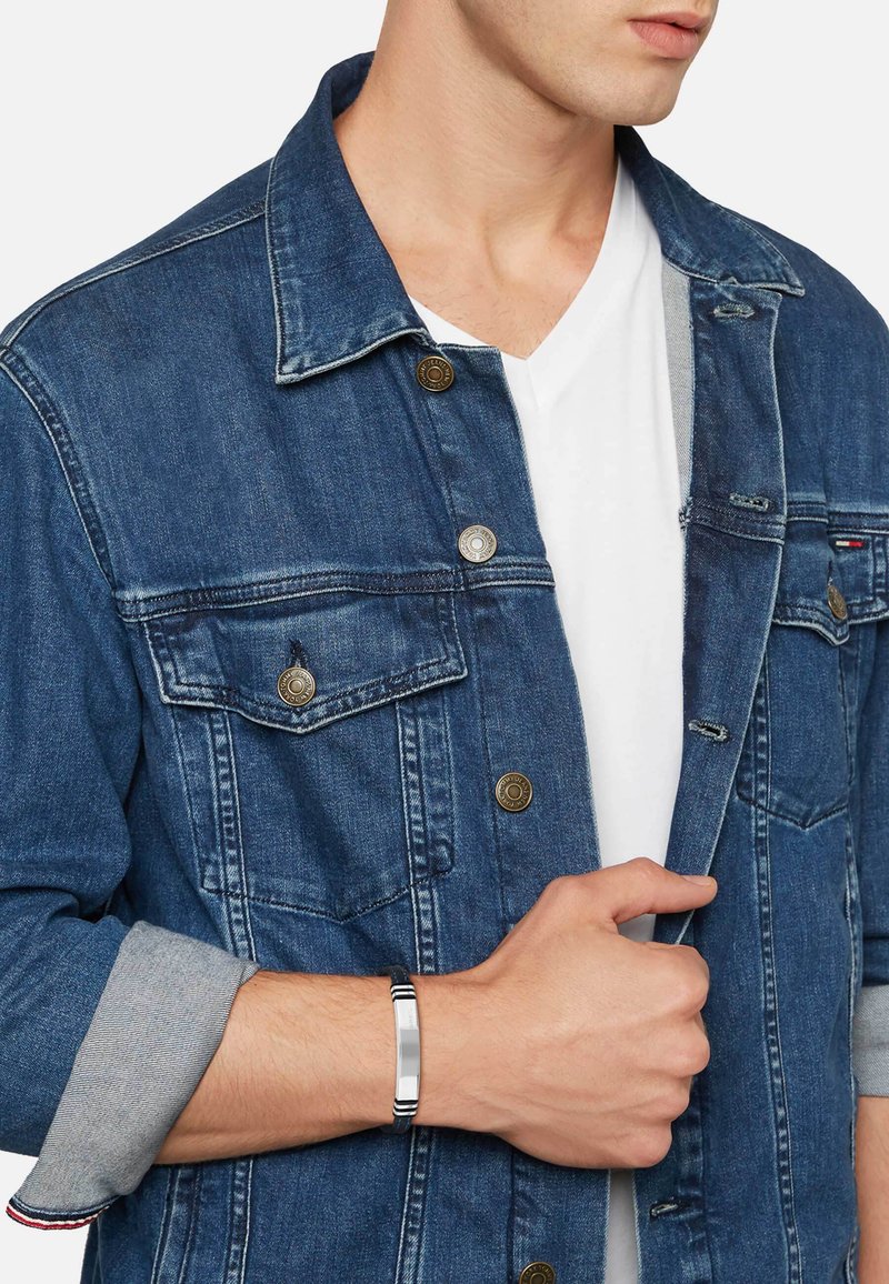 Man som bär en blå denimjacka över en vit V-ringad skjorta, håller i jackans krage med ena handen, bär ett silver- och svart armband.