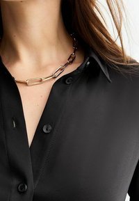 Musta nappipaita sileällä tekstuurilla, jossa on kullanvärinen ja tummanvärinen ketjulinkkikaulakoru, joka tuo kontrastia.