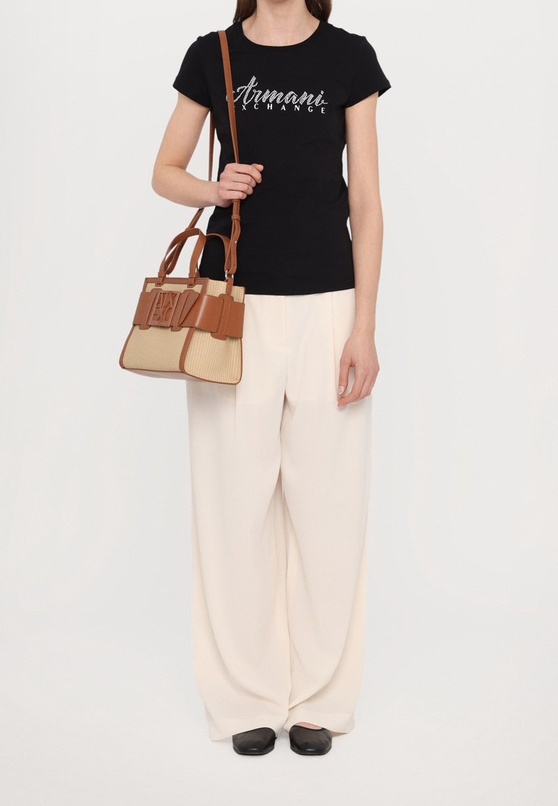Femme portant un t-shirt noir Armani Exchange et un pantalon blanc à jambes larges, tenant un sac à main en cuir beige et marron sur l'épaule.