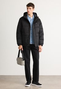 Jovem vestindo uma jaqueta puffer preta sobre uma camisa de ganga, calças pretas e ténis pretos, segurando uma pequena mala cruzada cinza.