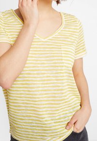 Geel en wit gestreepte V-hals T-shirt met korte mouwen, inclusief een linkerschouderzak, gemaakt van een lichtgewicht stof.