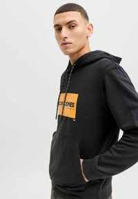 Jeune homme aux cheveux courts portant un sweat à capuche noir avec un logo rectangulaire orange et la main droite dans la poche avant.