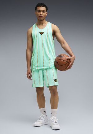 Young man in mint green basketball jersey and shorts holding a basketball, wearing white Puma sneakers and socks, standing against gray background.