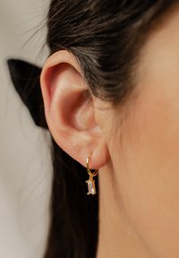 Orecchino a cerchio in oro con un rettangolo di pietra trasparente, fissato al cerchio con piccoli accenti metallici, indossato su un orecchio umano.