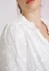 Weiße, bestickte Bluse mit floralen Mustern und feinem, rüschengeschmücktem Ausschnitt. Weiches, durchsichtiges Material mit puffärmel. Silberne Creolen sind sichtbar.