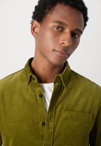 Cămașă verde din corduroy cu guler tip button-down, textură cu dungi verticale, nasturi negri și un buzunar pe piept. Purtați-o peste un tricou alb.