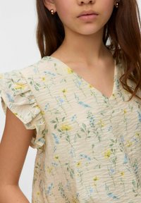 Top con motivo floreale in tessuto leggero e testurizzato. Presenta una scollatura a V e maniche a balze, con fiori gialli e blu su uno sfondo crema.