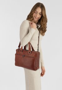 Borsa a mano in pelle marrone con forma rettangolare, doppio manico superiore, tracolla staccabile e tasca frontale con zip visibile.