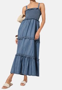 Denim maxiklänning med smockad byst, rufsiga axelremmar och lager-på-lager kjol. Färgen är medelblå med en mjuk textur. Accessoarer inkluderar en vävd väska.