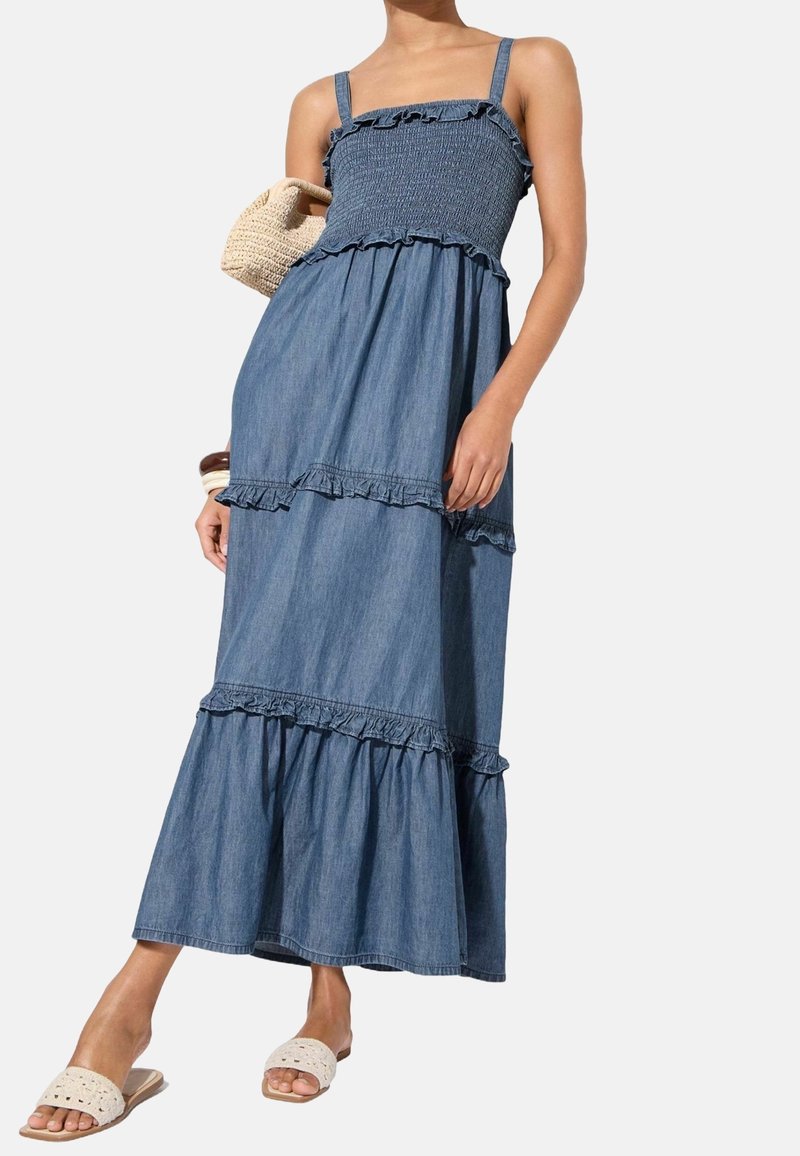 Denim maxiklänning med smockad byst, rufsiga axelremmar och lager-på-lager kjol. Färgen är medelblå med en mjuk textur. Accessoarer inkluderar en vävd väska.